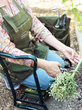 woman wearing green apron kneels on garden kneeler and holds pruner