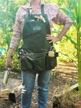 woman wearing green apron with pockets holds trowel in vegetable garden background